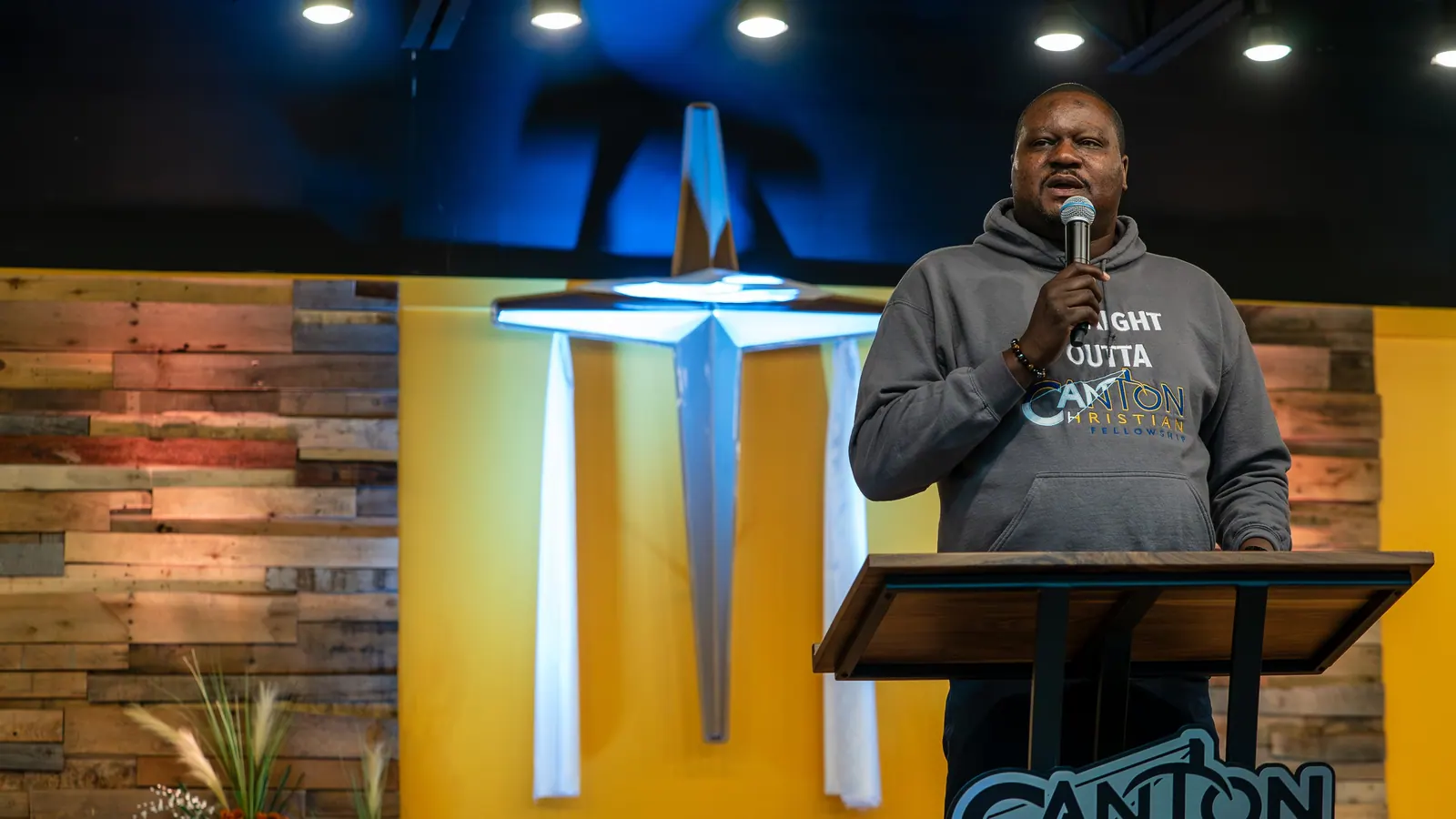 Pastor Washington preaching at the pulpit, the lit cross and wood wall behind him